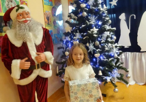 Uśmiechnięta dziewczynka trzyma w ręku prezent, stoi obok figury Mikołaja.